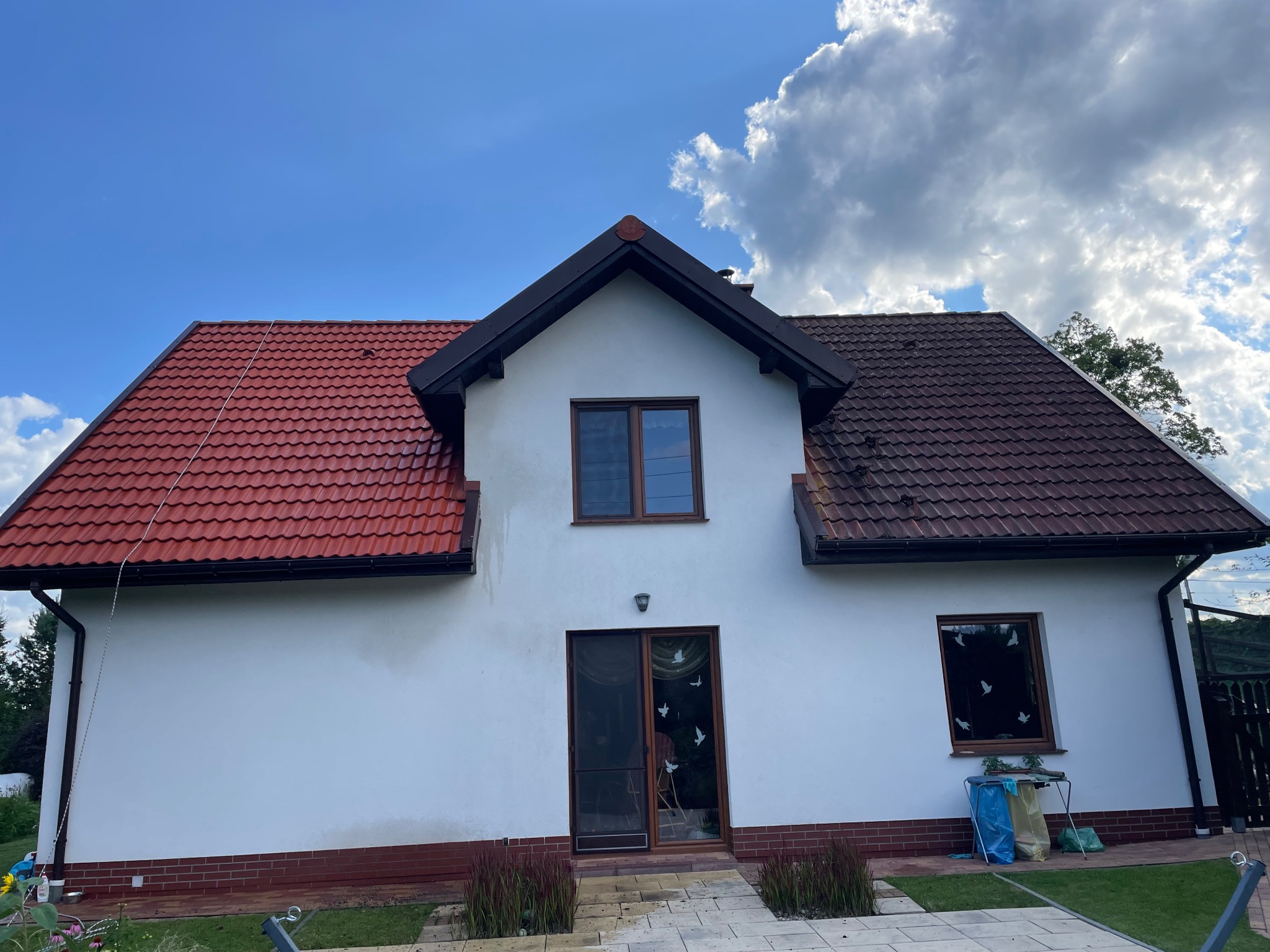 Dom z dachem częściowo wyczyszczonym, ukazując różnicę w kolorze i czystości połaci. Widoczne białe ściany i fragment kostki brukowej. Niebo z chmurami w tle.
