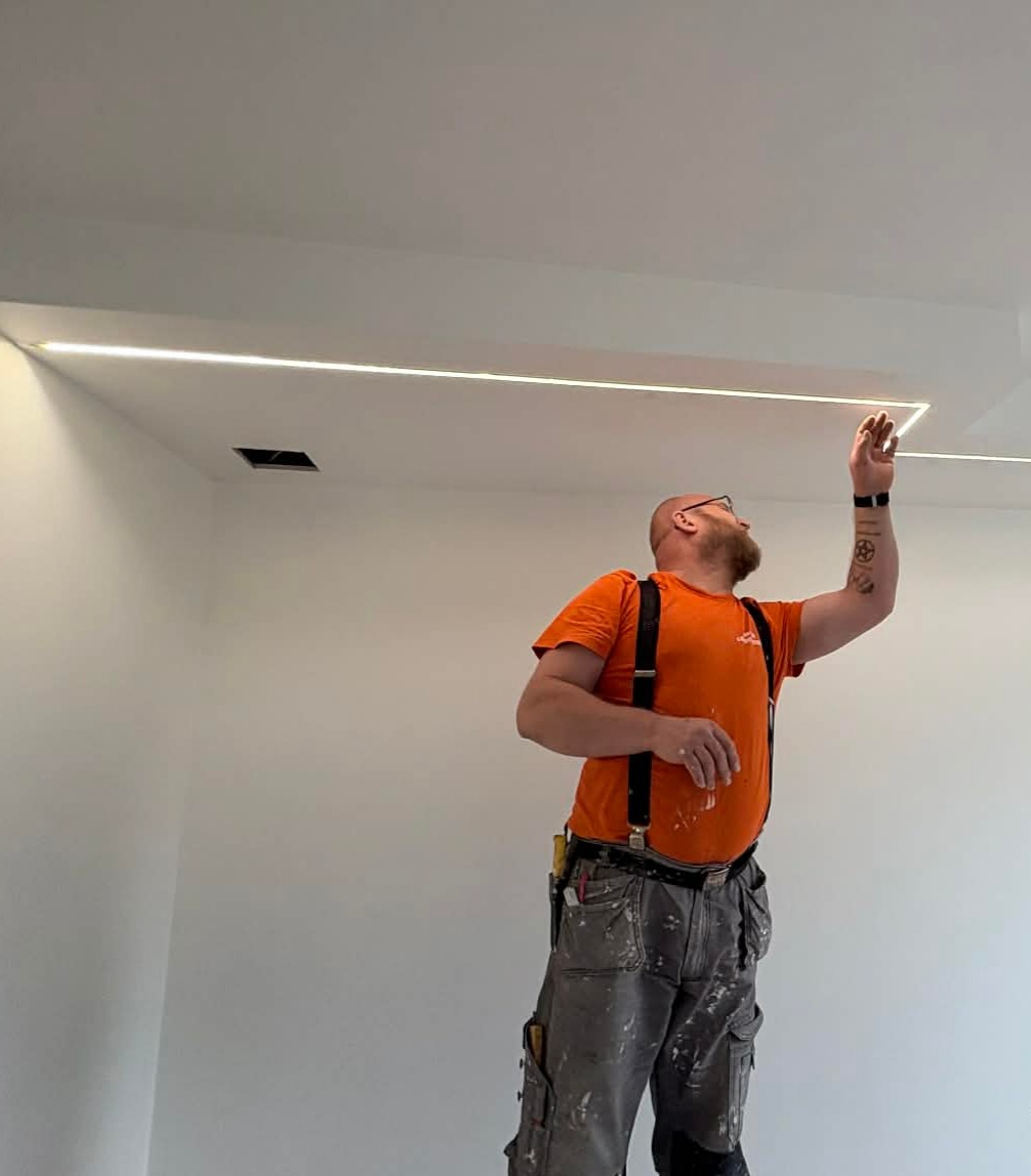 Pracownik na drabinie montuje oświetlenie LED w suficie podwieszanym. Widoczne narzędzia i ubranie robocze z plamami farby. Białe ściany i sufit.