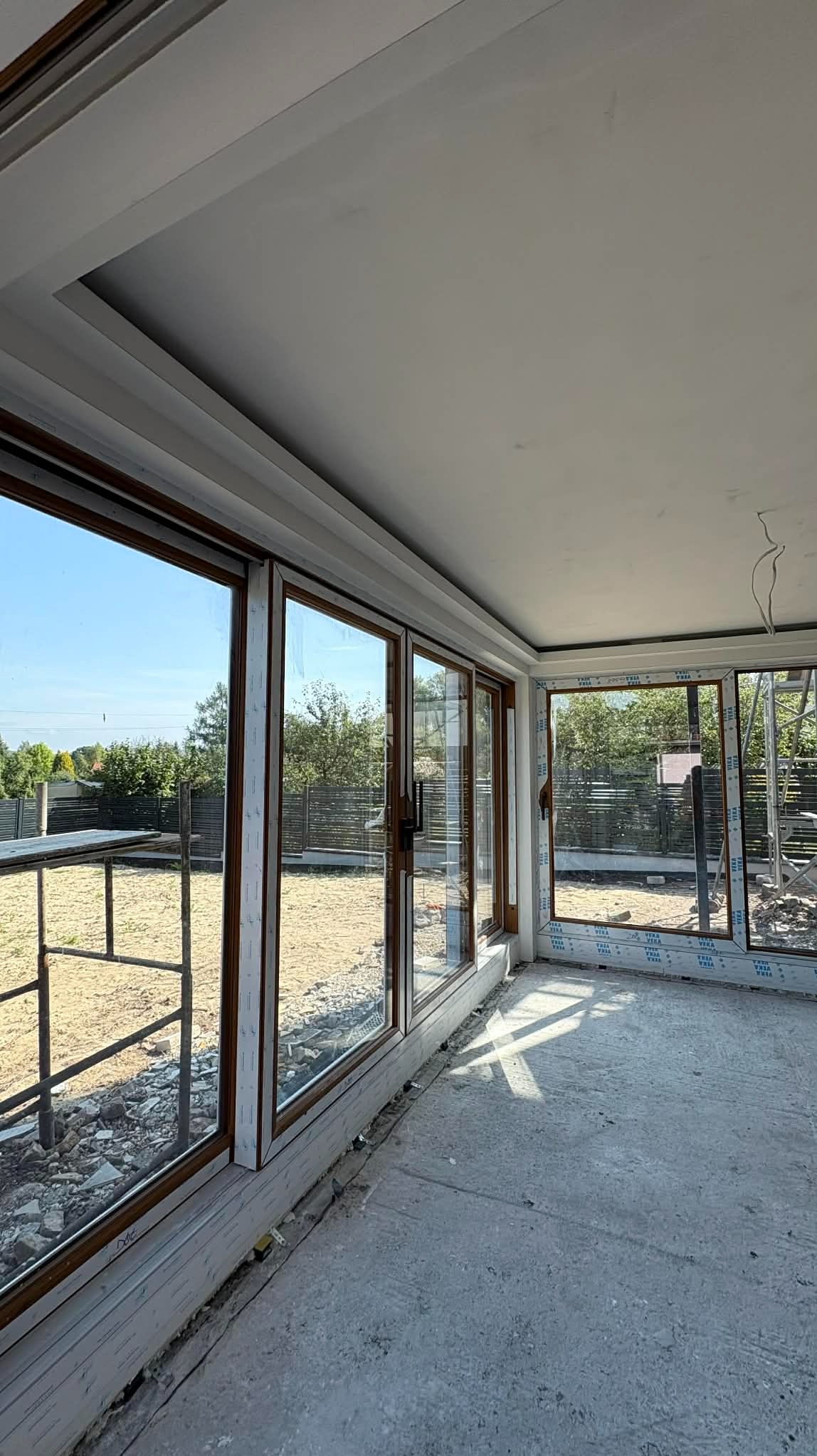 Wnętrze w trakcie wykańczania z dużymi oknami przesuwnymi z brązowymi ramami. Widoczne surowe ściany i podłoga, przygotowane do dalszych prac wykończeniowych.