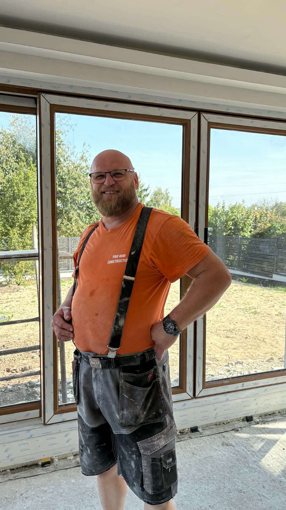 Uśmiechnięty monter w szelkach, na tle okien z widokiem na ogród. Widoczne narzędzia i elementy montażowe. Jasne wnętrze w trakcie prac wykończeniowych.