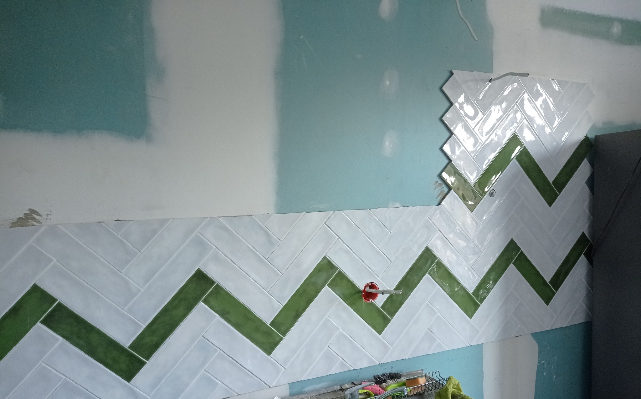 Ściana w trakcie wykańczania z białymi i zielonymi płytkami ułożonymi w jodełkę. Widoczne niedokończone fragmenty i instalacja elektryczna.