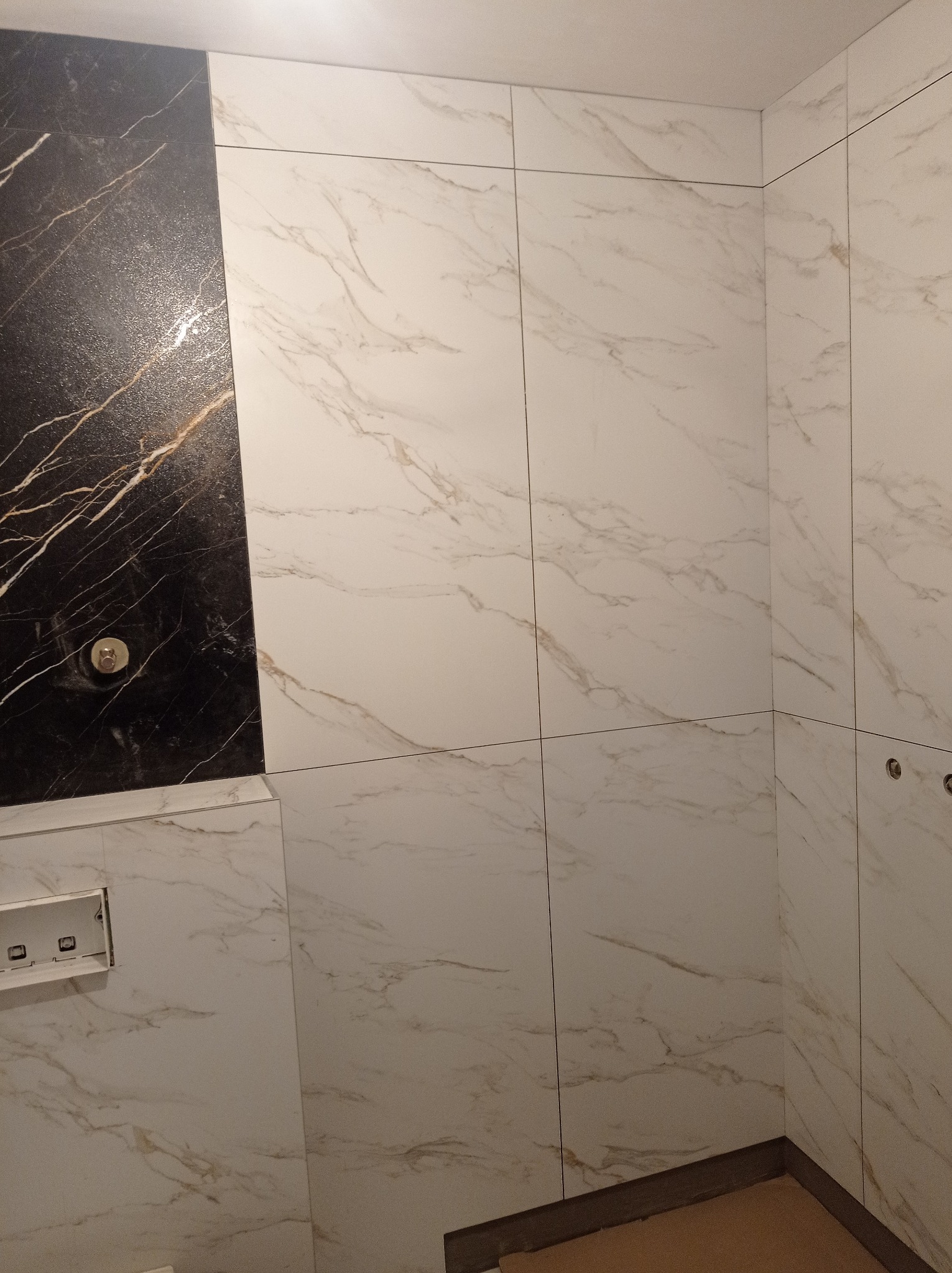 Ściana wykończona płytkami imitującymi marmur w odcieniach bieli i czerni z widocznymi otworami na instalacje. Ułożenie płytek w łazience.