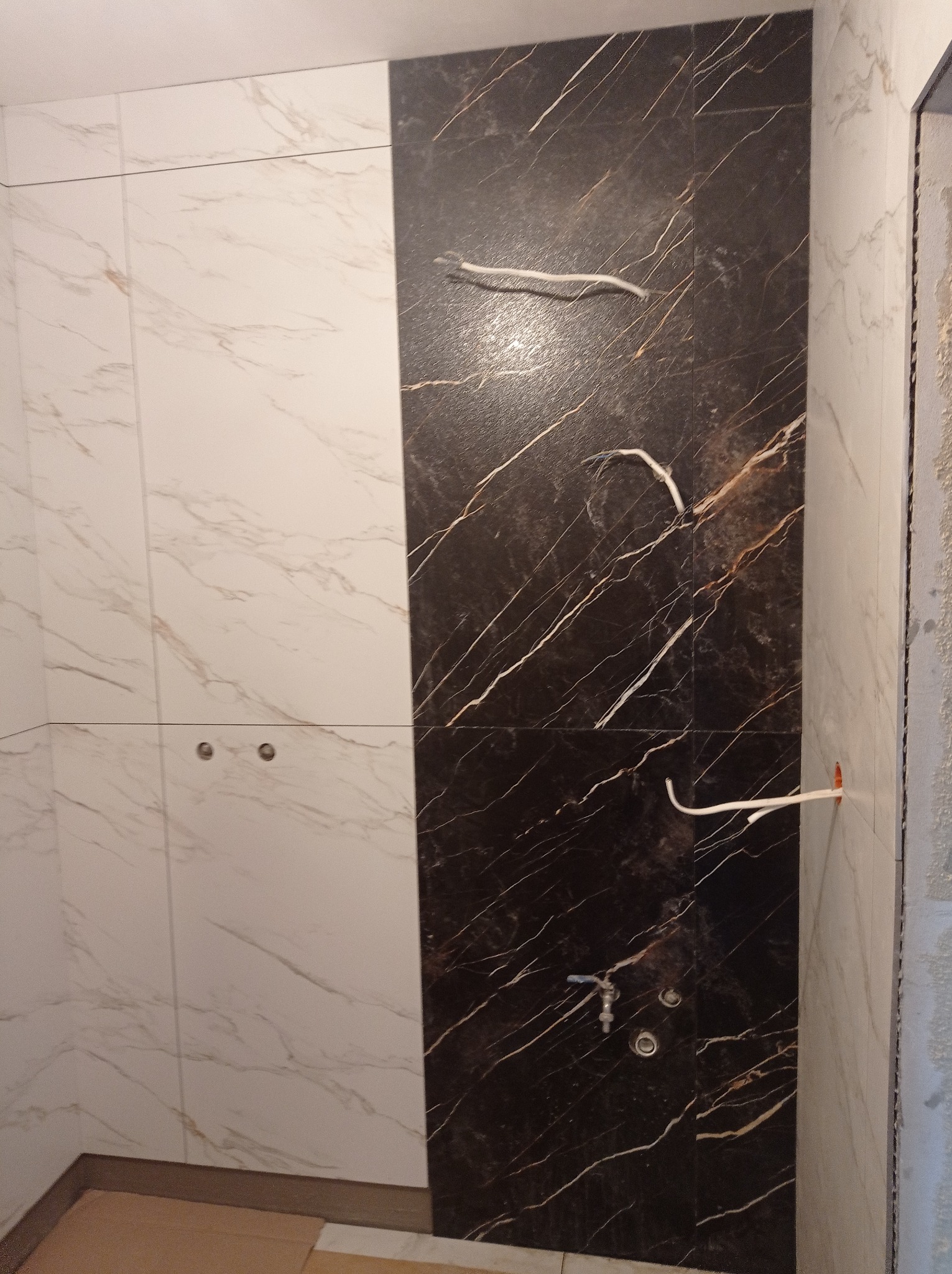 Ściana w łazience wyłożona płytkami imitującymi marmur w odcieniach bieli i brązu. Widoczne wystające kable i rurka doprowadzająca wodę. Fragment ściany surowej.