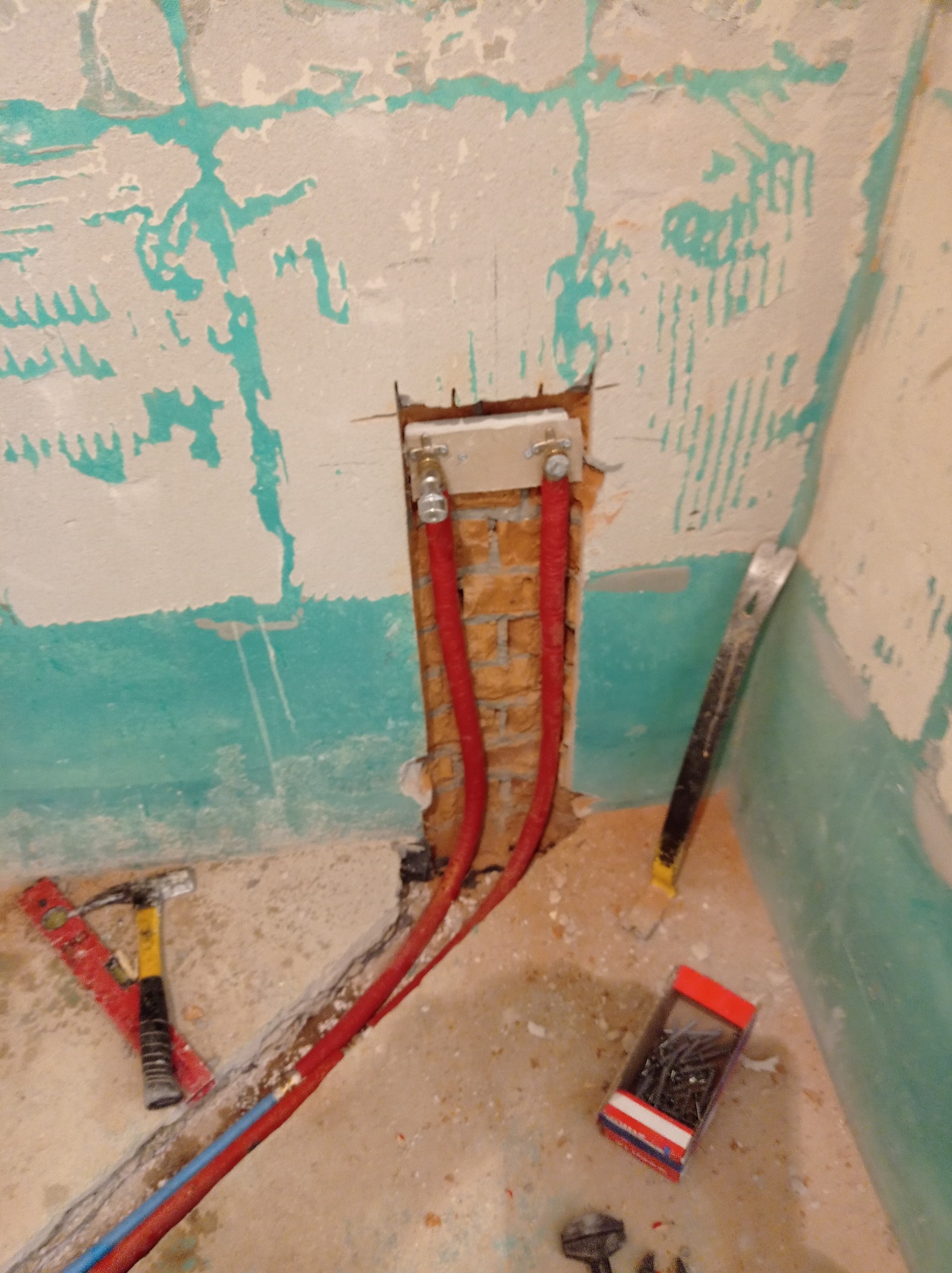 Montaż instalacji wodnej w łazience: czerwone rury prowadzone w ścianie z cegły, narzędzia glazurnicze na podłodze. Ściana w trakcie remontu, z resztkami farby.