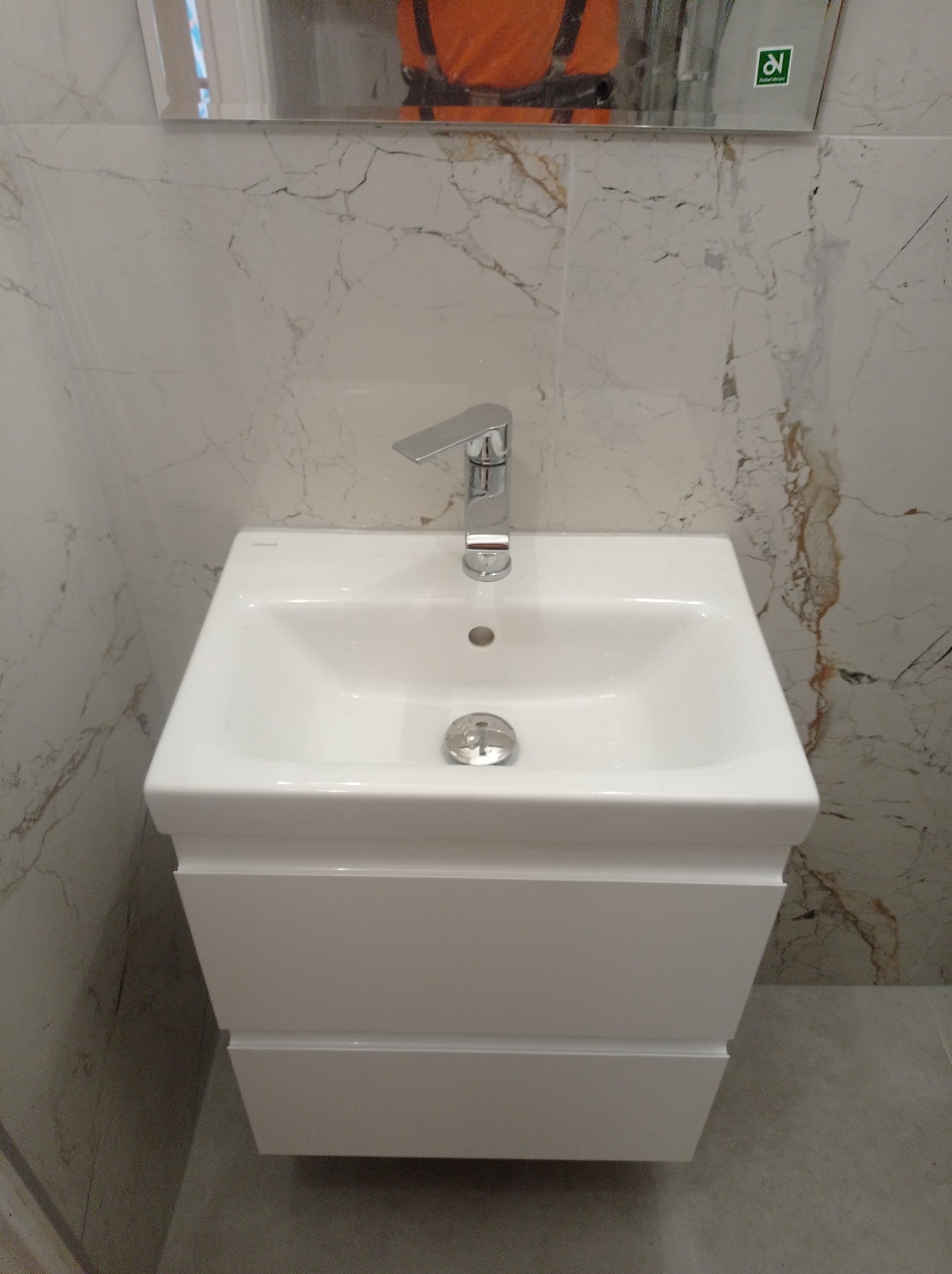 Biała umywalka z szafką w łazience wykończonej marmurowymi płytkami. Bateria chromowana, odbicie osoby w lustrze. Minimalistyczny design.