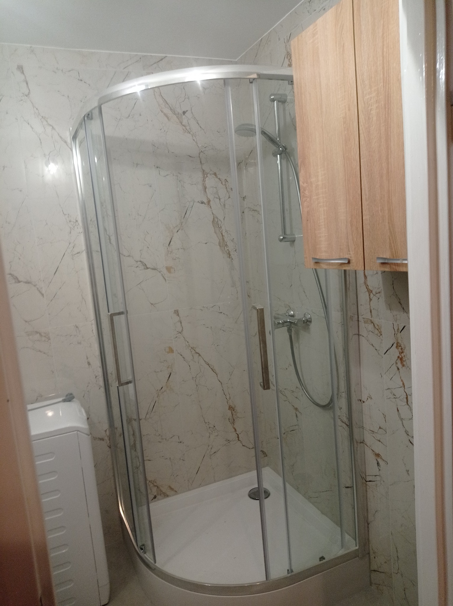 Nowoczesna kabina prysznicowa z marmurowymi płytkami i drewnianą szafką w łazience. Widoczna pralka z boku. Ujęcie z bliska, prezentujące detale wykończenia.