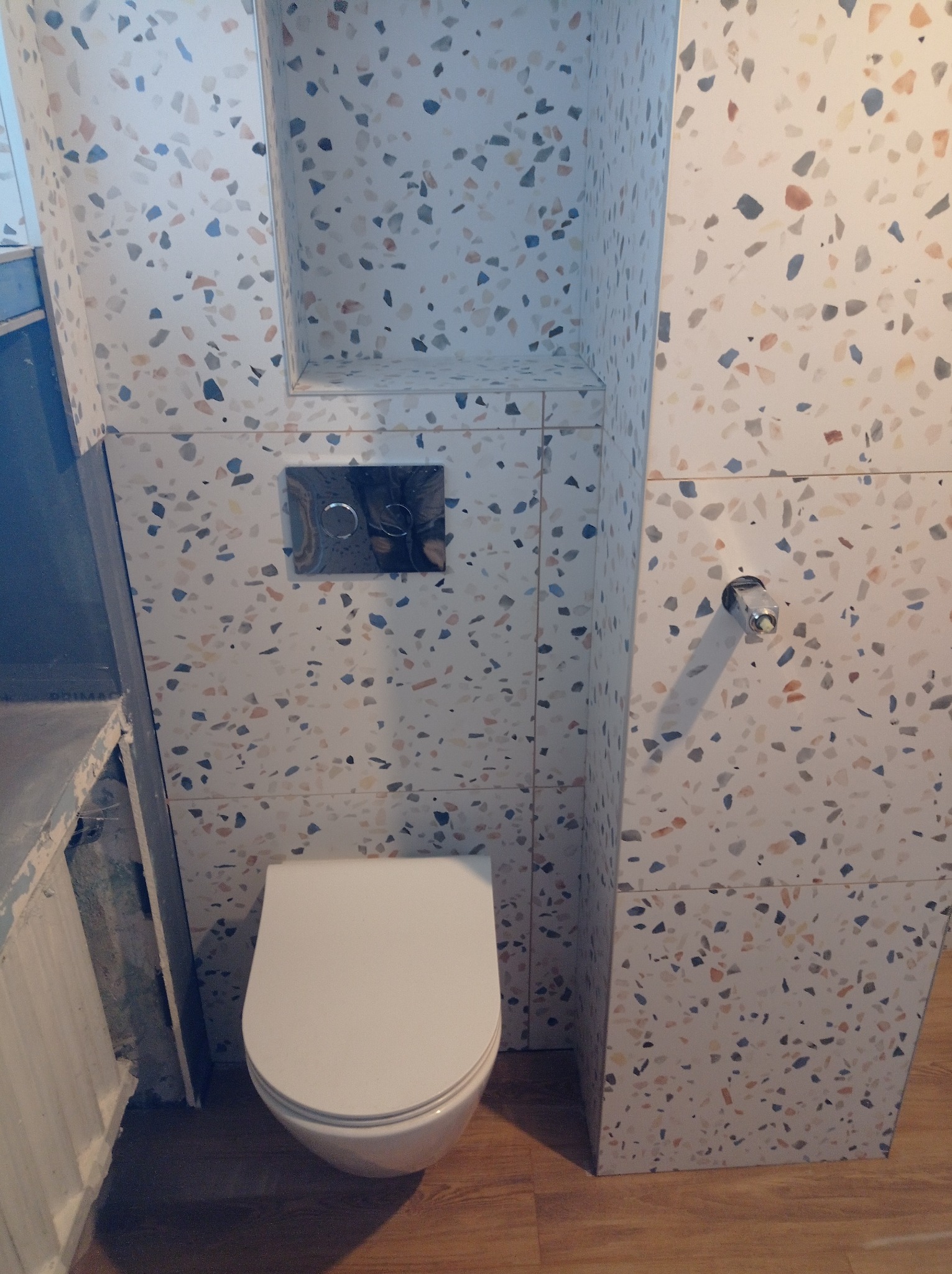 Nowoczesna toaleta w łazience wykończonej płytkami w stylu lastryko. Widoczny stelaż podtynkowy i drewnopodobna podłoga. Minimalistyczny design.