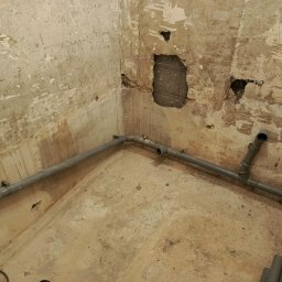 Marcin Szewczuk - Surowe wnętrze z szarymi rurami kanalizacyjnymi w narożniku, ściany z widocznymi śladami po kuciu, przygotowane do dalszych prac wykończeniowych.