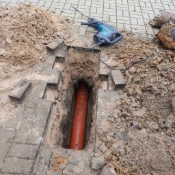 Marcin Szewczuk - Pomarańczowa rura w wykopie z kostki brukowej, obok leży młot udarowy i ziemia. Widoczne fragmenty kostki brukowej i narzędzia sugerują prace instalacyjne.