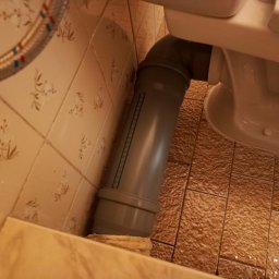 Marcin Szewczuk - Szara rura kanalizacyjna odpływowa podłączona do toalety na tle beżowych płytek z wzorem kwiatowym i brązowych płytek podłogowych.