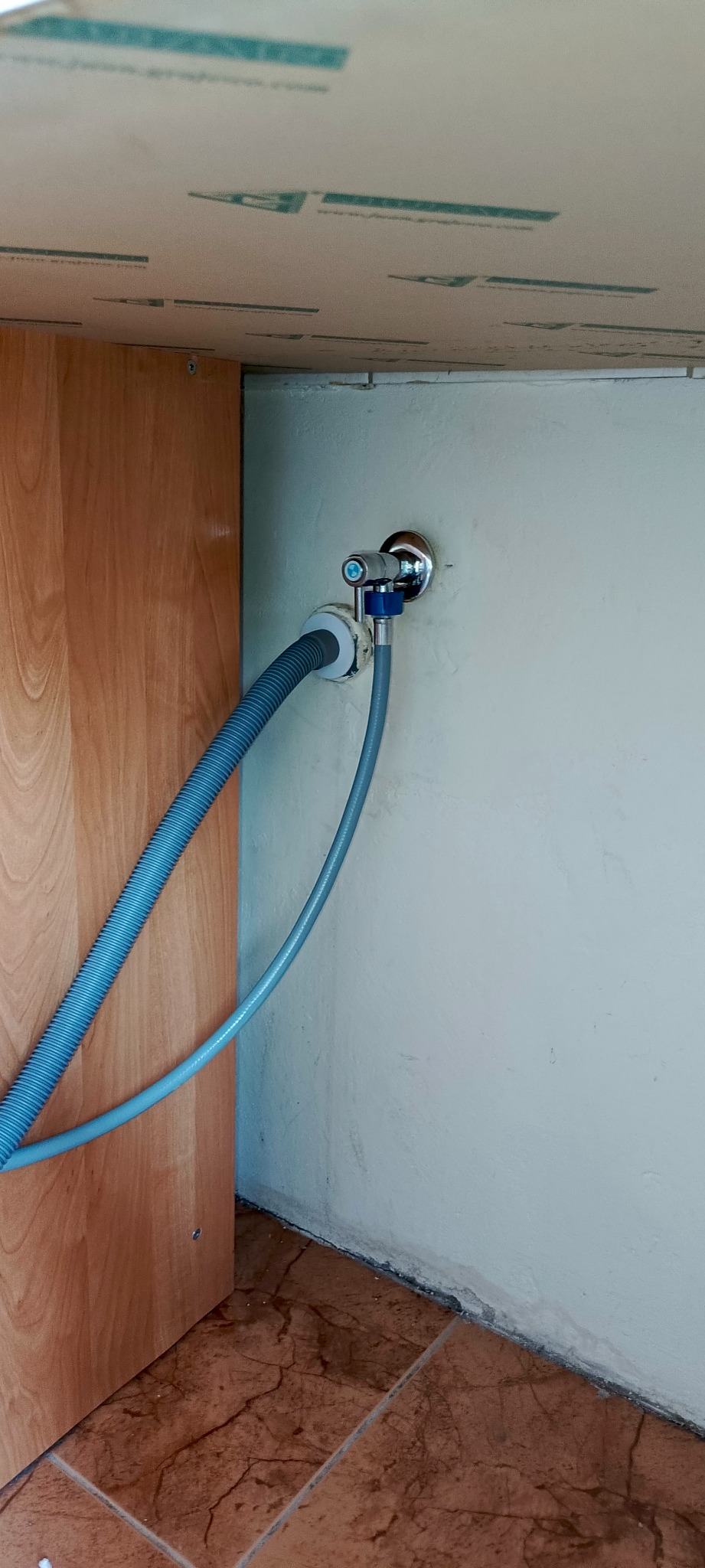 Podłączenie węża do zaworu pod umywalką/zlewozmywakiem. Widoczny chromowany zawór, szary wąż karbowany oraz gładki, fragment szafki i brązowe płytki podłogowe.