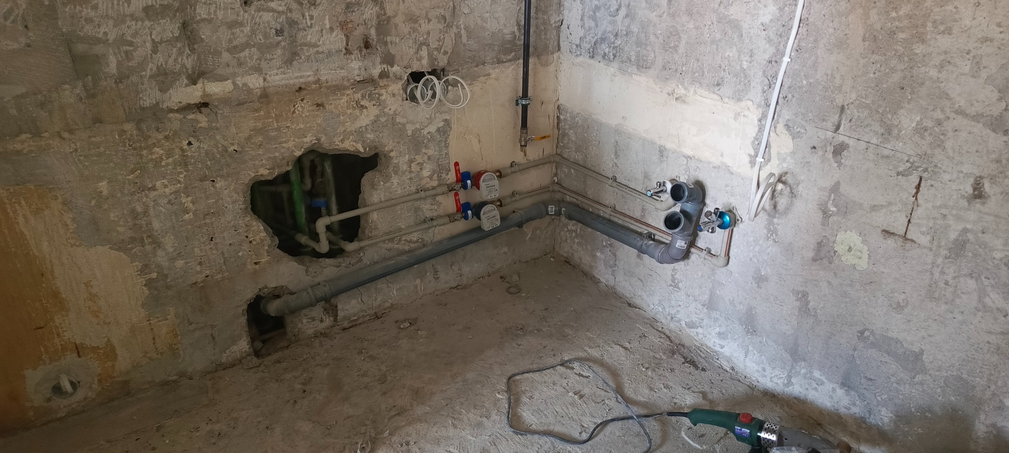 Surowy stan pomieszczenia z częściowo wykonaną instalacją hydrauliczną: rury, zawory, liczniki. W tle ściany w stanie deweloperskim, widoczne narzędzie do spawania rur.