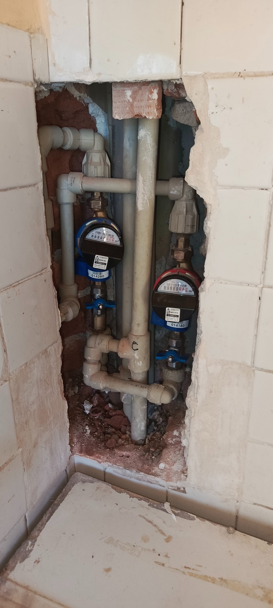Widok na instalację wodną z dwoma licznikami, ukrytą w ścianie z fragmentami uszkodzonych płytek. Rury i liczniki w jasnych barwach. Prace hydrauliczne.