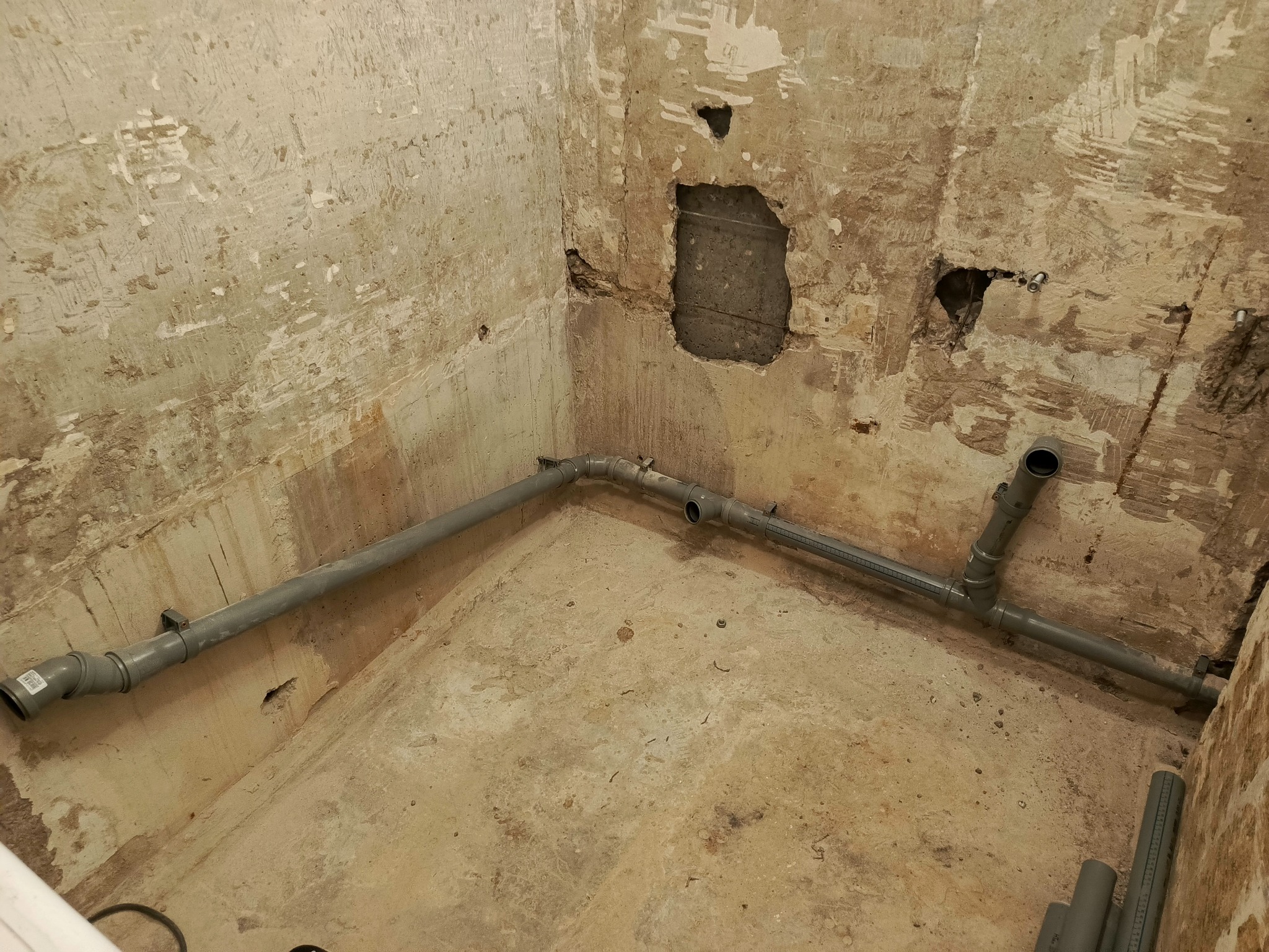 Surowe wnętrze z szarymi rurami kanalizacyjnymi w narożniku, ściany z widocznymi śladami po kuciu, przygotowane do dalszych prac wykończeniowych.