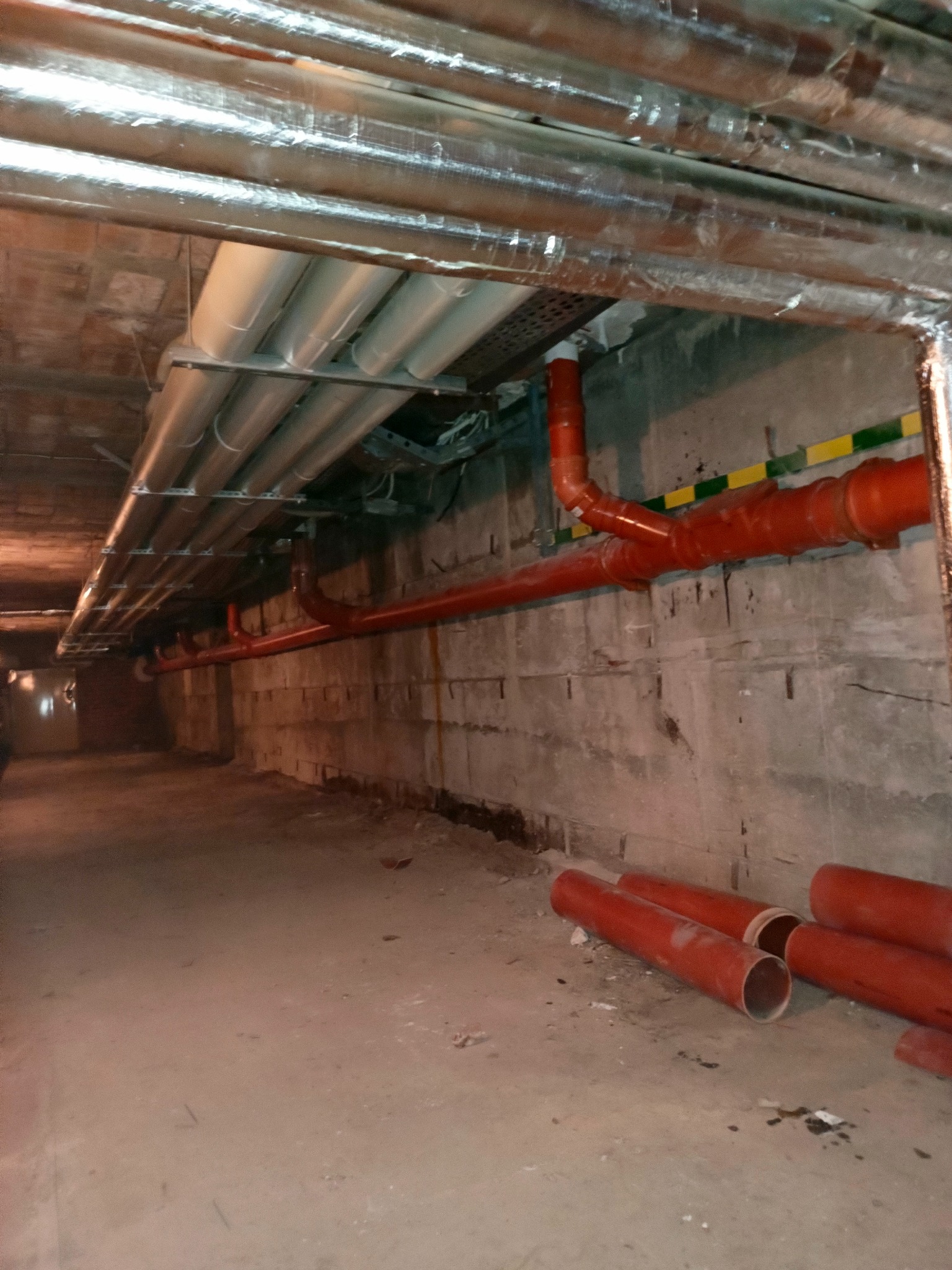 Instalacja rur w piwnicy: czerwone rury kanalizacyjne ułożone na podłodze i pod stropem, srebrne rury wentylacyjne pod sufitem. Widoczne łączenia i mocowania.
