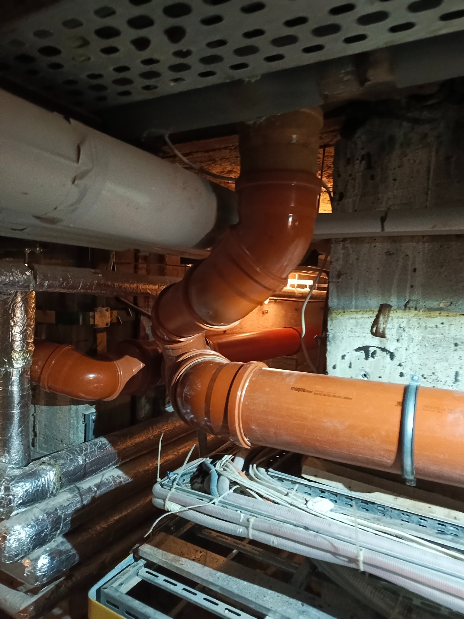 Pomarańczowe rury kanalizacyjne z tworzywa sztucznego, zamontowane pod sufitem w piwnicy. Widoczne elementy instalacji i kable. Niska perspektywa.