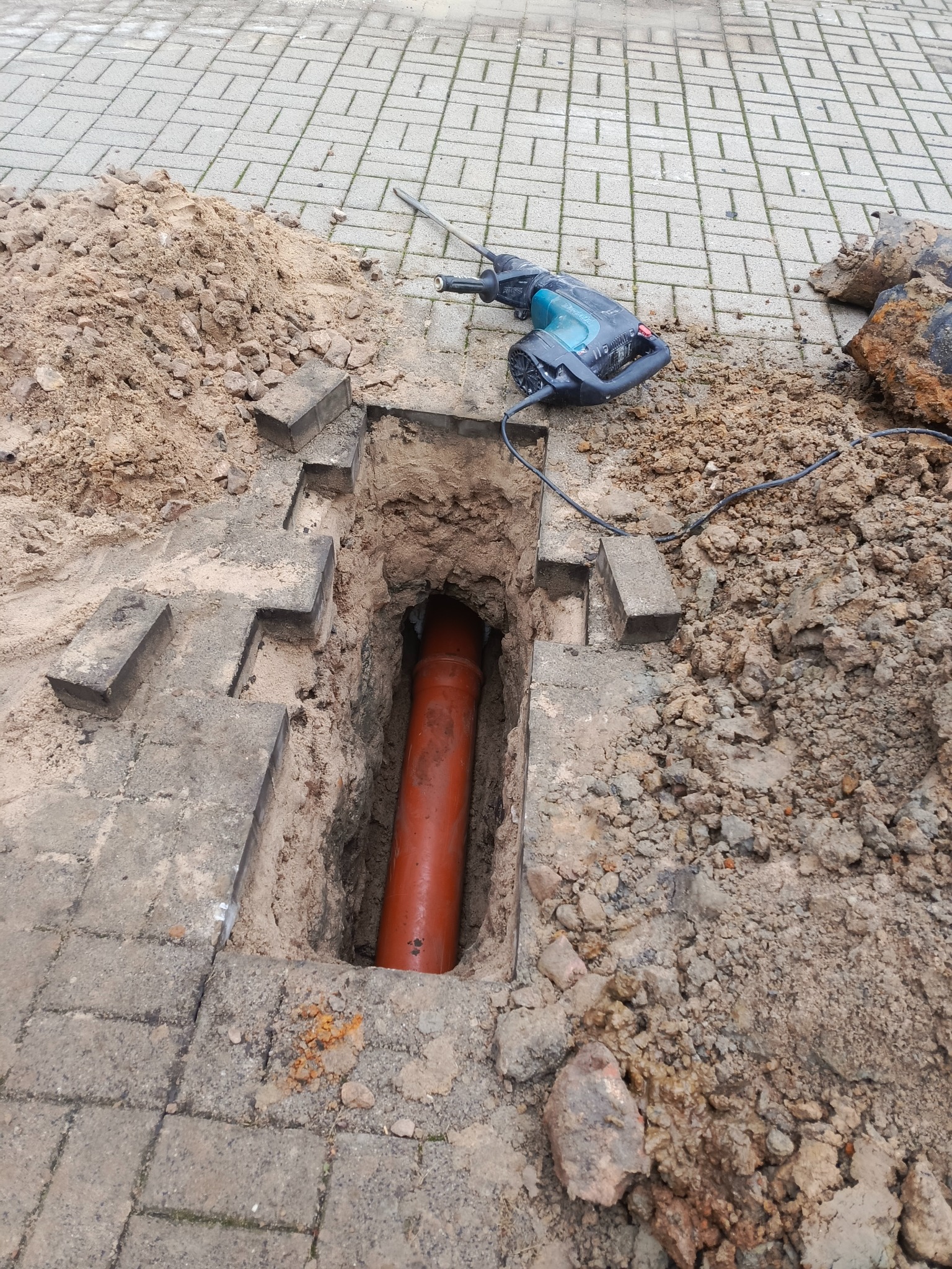 Pomarańczowa rura w wykopie z kostki brukowej, obok leży młot udarowy i ziemia. Widoczne fragmenty kostki brukowej i narzędzia sugerują prace instalacyjne.