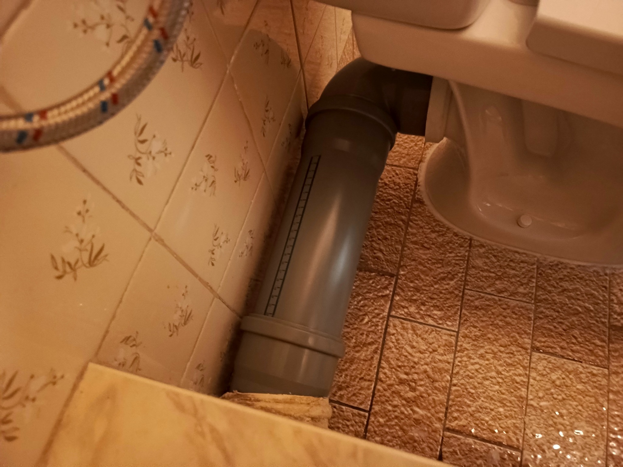 Szara rura kanalizacyjna odpływowa podłączona do toalety na tle beżowych płytek z wzorem kwiatowym i brązowych płytek podłogowych.