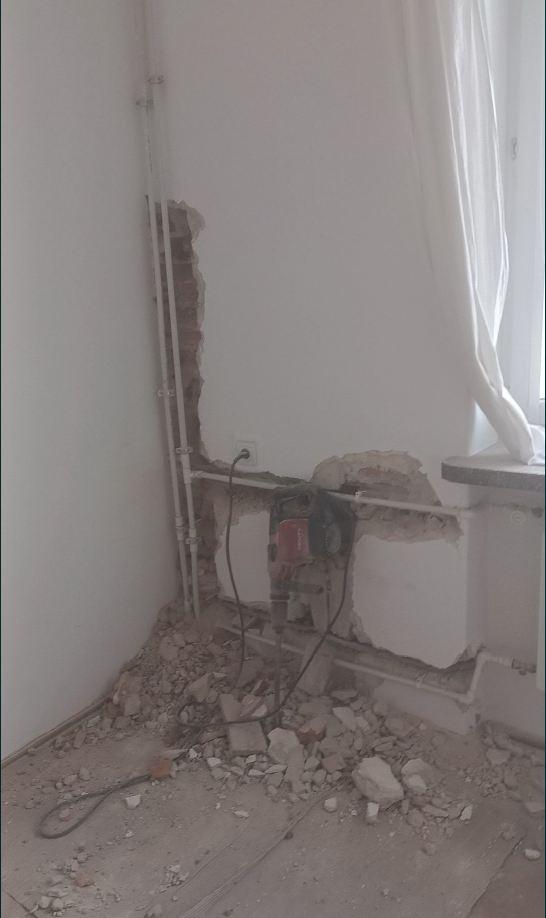 Demontaż fragmentu ściany pod instalacje, widoczne rury i gruz. W tle okno z białą zasłoną. Użycie wiertarki udarowej.