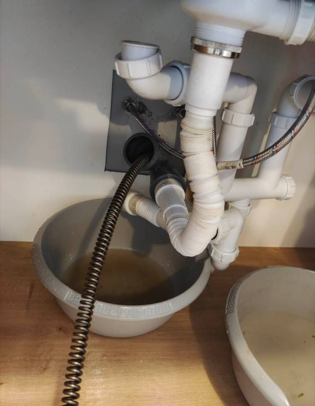 Udrażnianie odpływu zlewu spiralą hydrauliczną; woda zbierana do miski. Widoczne białe rury odpływowe i szara płyta montażowa. Praca w trakcie.