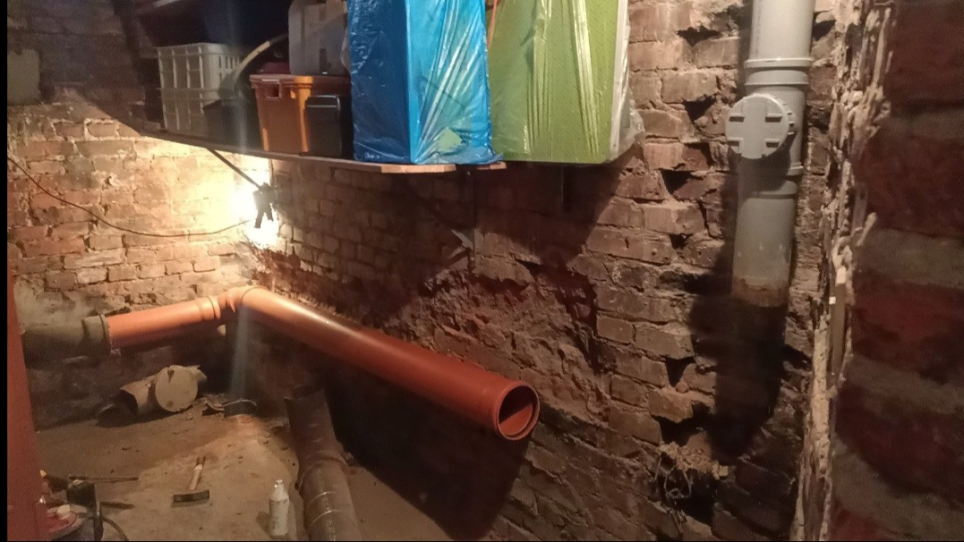 Pomarańczowe rury kanalizacyjne w piwnicy z ceglanymi ścianami. Widoczne też szare rury i narzędzia. Na półce plastikowe pojemniki.