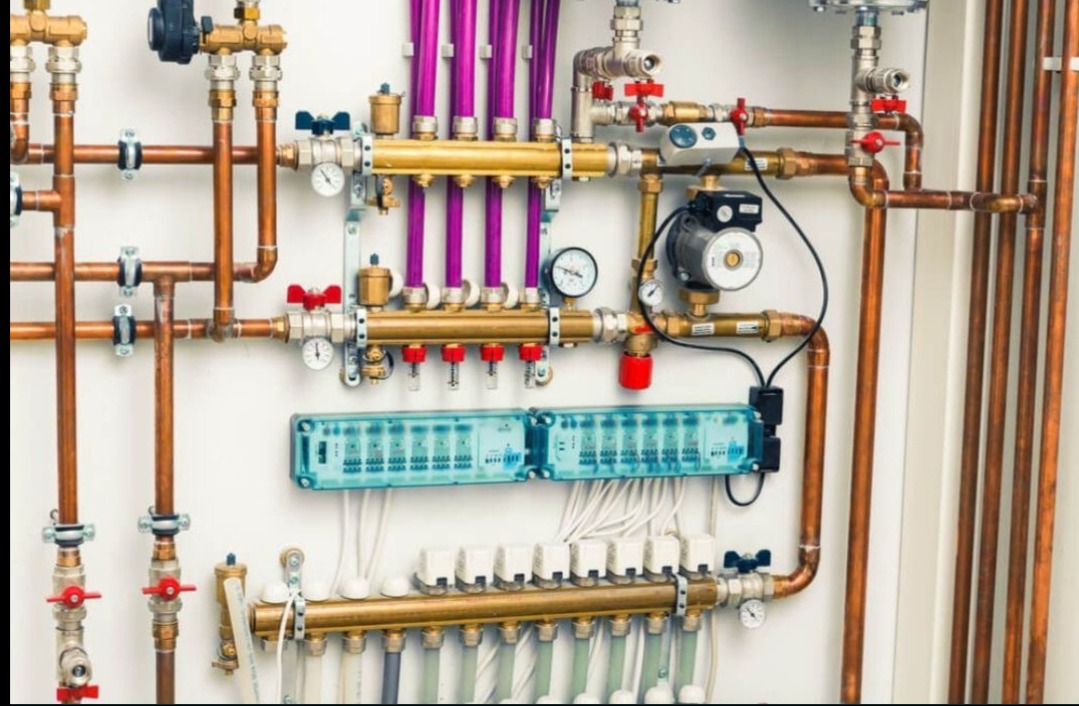 Rozbudowany system instalacji hydraulicznej z miedzianymi rurami, zaworami i rozdzielaczami na białej ścianie. Widoczne manometry i elementy automatyki.