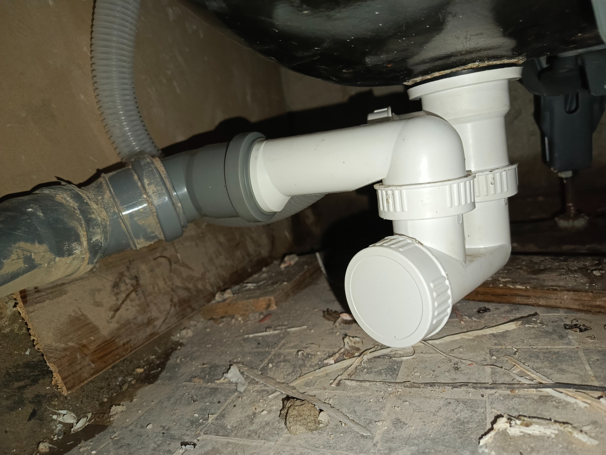 Szare i białe rury odpływowe pod zlewem, z widocznymi złączkami i elementami instalacji. Widoczny fragment podłogi z zabrudzeniami i resztkami materiałów budowlanych.