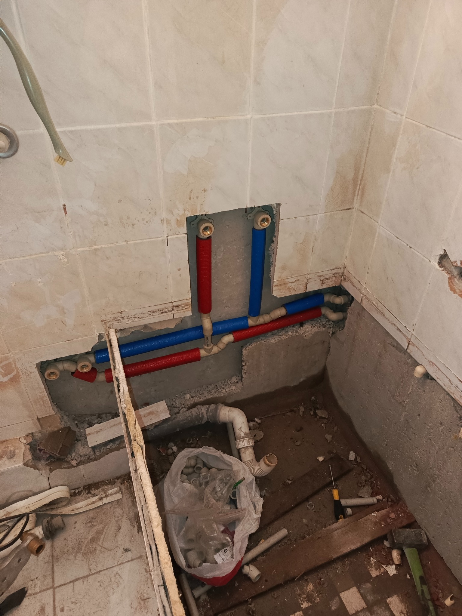 Instalacja rur wodnych (czerwone i niebieskie) w ścianie z płytek, widoczne narzędzia i akcesoria hydrauliczne w trakcie prac wykończeniowych.