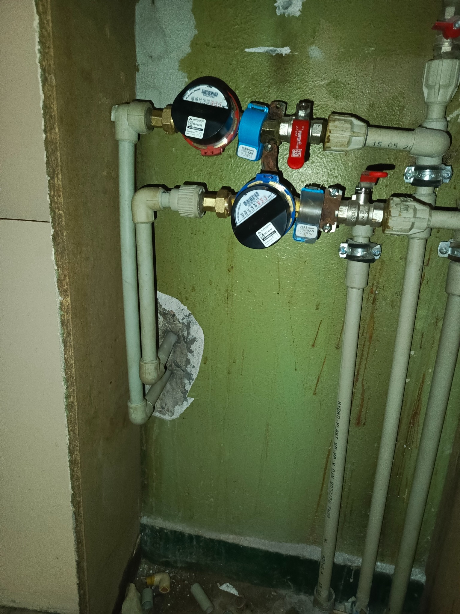 Zamontowane wodomierze z zaworami i rurami z tworzywa sztucznego na zielonej ścianie. Widoczne ślady po wcześniejszych pracach instalacyjnych i fragment szafki.
