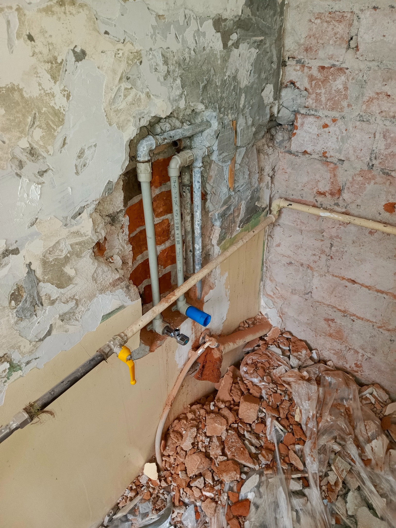 Fragment ściany z odsłoniętą instalacją hydrauliczną w trakcie remontu. Widoczne rury, zawory i gruz ceglany. Ściana w trakcie skuwania tynku.