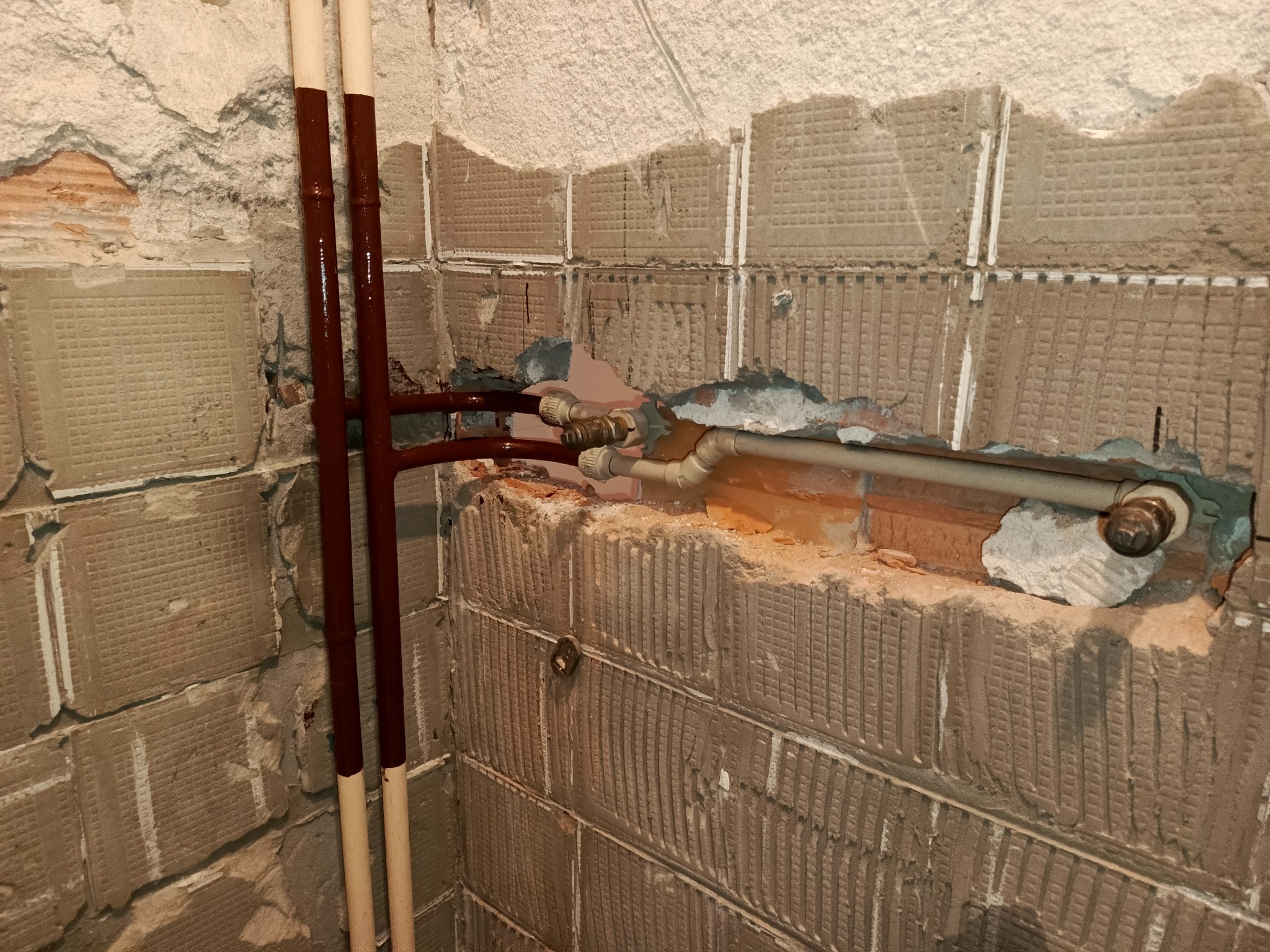 Odkryte rury wodne i grzewcze w ścianie z resztkami płytek, widoczne łączenia i zawory, ściana w trakcie remontu, instalacja w trakcie przeróbki.