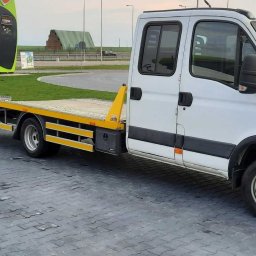 Sprowadzanie aut, holowanie - Biała laweta Iveco z żółtymi elementami, zaparkowana na kostce brukowej, w tle stacja benzynowa i pole. Widoczne tablice informacyjne i zarysy zabudowań.