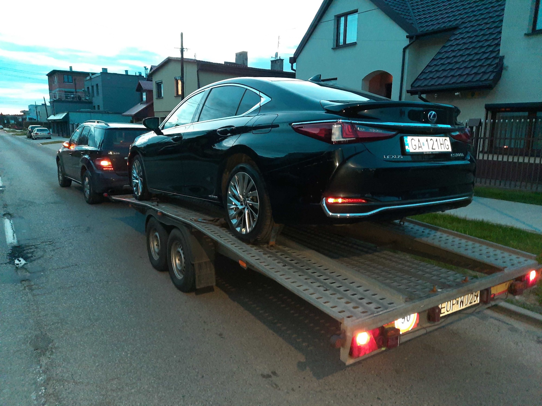 Czarny Lexus ES 300h na ażurowej lawecie, holowany przez ciemny SUV na tle zabudowy jednorodzinnej. Widoczny numer rejestracyjny lawety i auta.