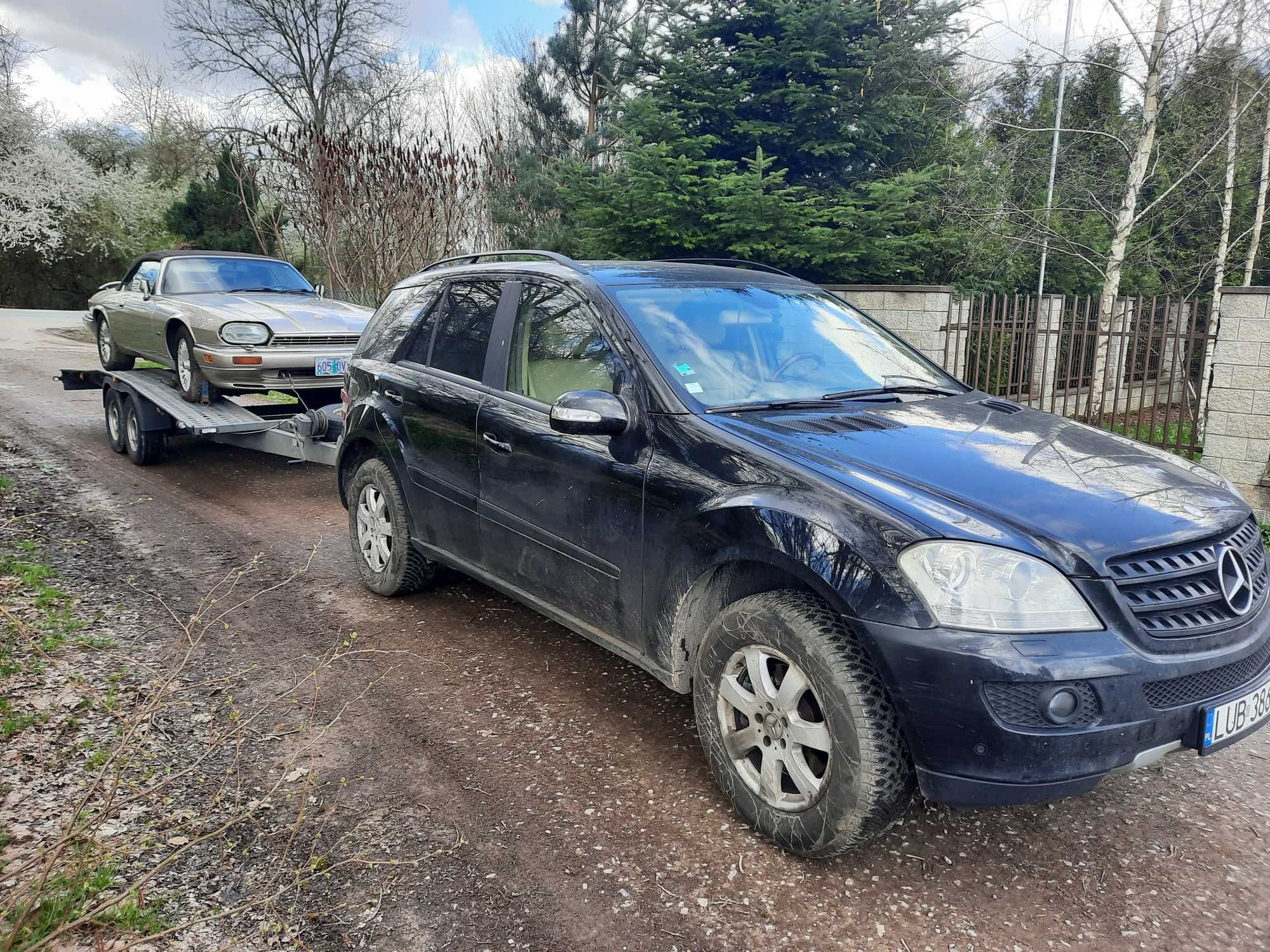 Czarny SUV Mercedes ciągnie lawetę z beżowym Jaguarem XJS cabrio na tle drzew i ogrodzenia. Widoczny błotnisty teren i tablica rejestracyjna.