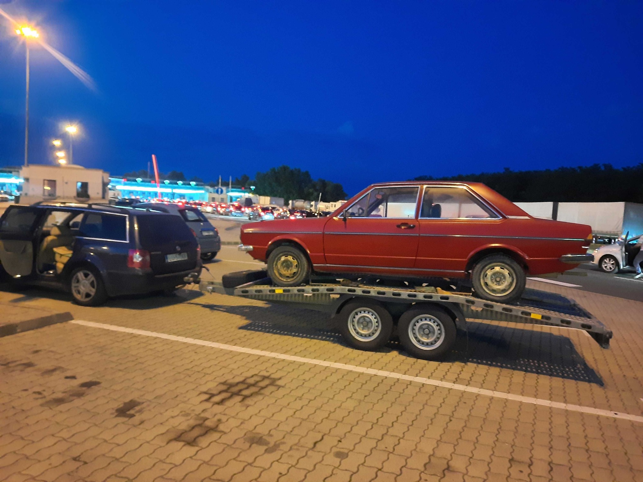 Czerwony samochód osobowy na lawecie, holowany przez granatowe kombi. Na parkingu, w tle budynki i inne pojazdy. Wieczorny transport pojazdu.