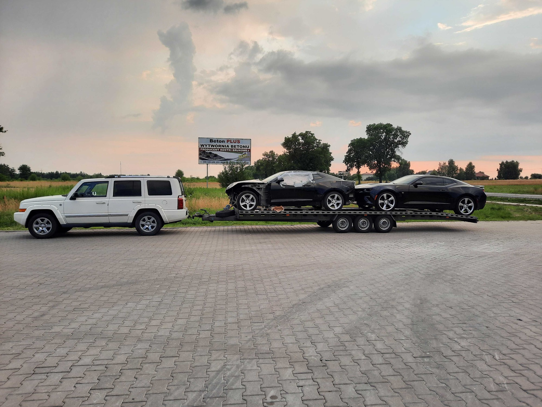 Biały Jeep Commander ciągnie lawetę z dwoma czarnymi Chevrolet Camaro, jeden uszkodzony, na tle billboardu i pochmurnego nieba o zmierzchu.