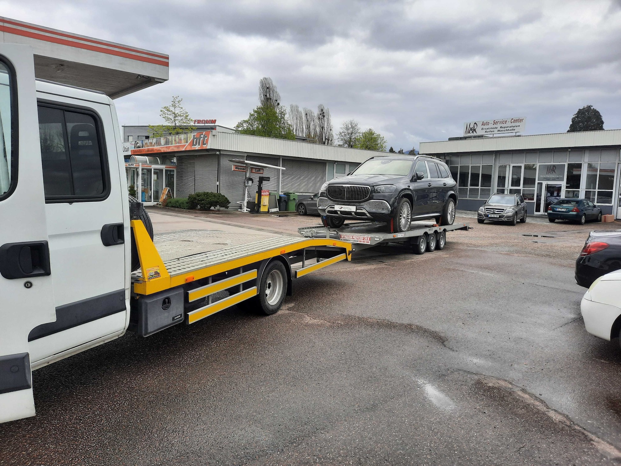 Czarny SUV na lawecie przed stacją paliw i warsztatem samochodowym. Laweta z żółtymi elementami, mokry asfalt. Pochmurne niebo.