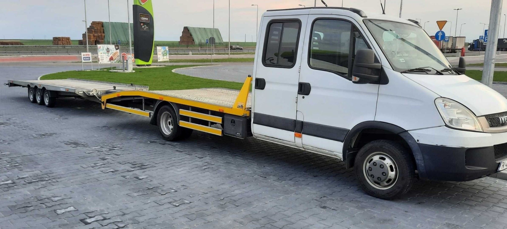 Biała laweta Iveco z żółtymi elementami, zaparkowana na kostce brukowej, w tle stacja benzynowa i pole. Widoczne tablice informacyjne i zarysy zabudowań.