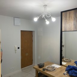 Ivan Sydorchuk - Jasne wnętrze po remoncie: białe ściany, marmurowe płytki podłogowe, drewniane drzwi i nowoczesna lampa sufitowa. Na stole narzędzia i kable.