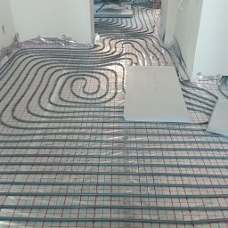 Ivan Sydorchuk - Szare rury ogrzewania podłogowego ułożone na folii z czerwoną siatką, widoczne w surowym wnętrzu. W tle zwój rur i fragmenty ścian.