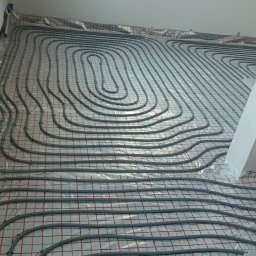 Ivan Sydorchuk - Szare rury ogrzewania podłogowego ułożone spiralnie na folii z czerwoną kratką, widoczne przy ścianie. W tle okno z różnymi przedmiotami.