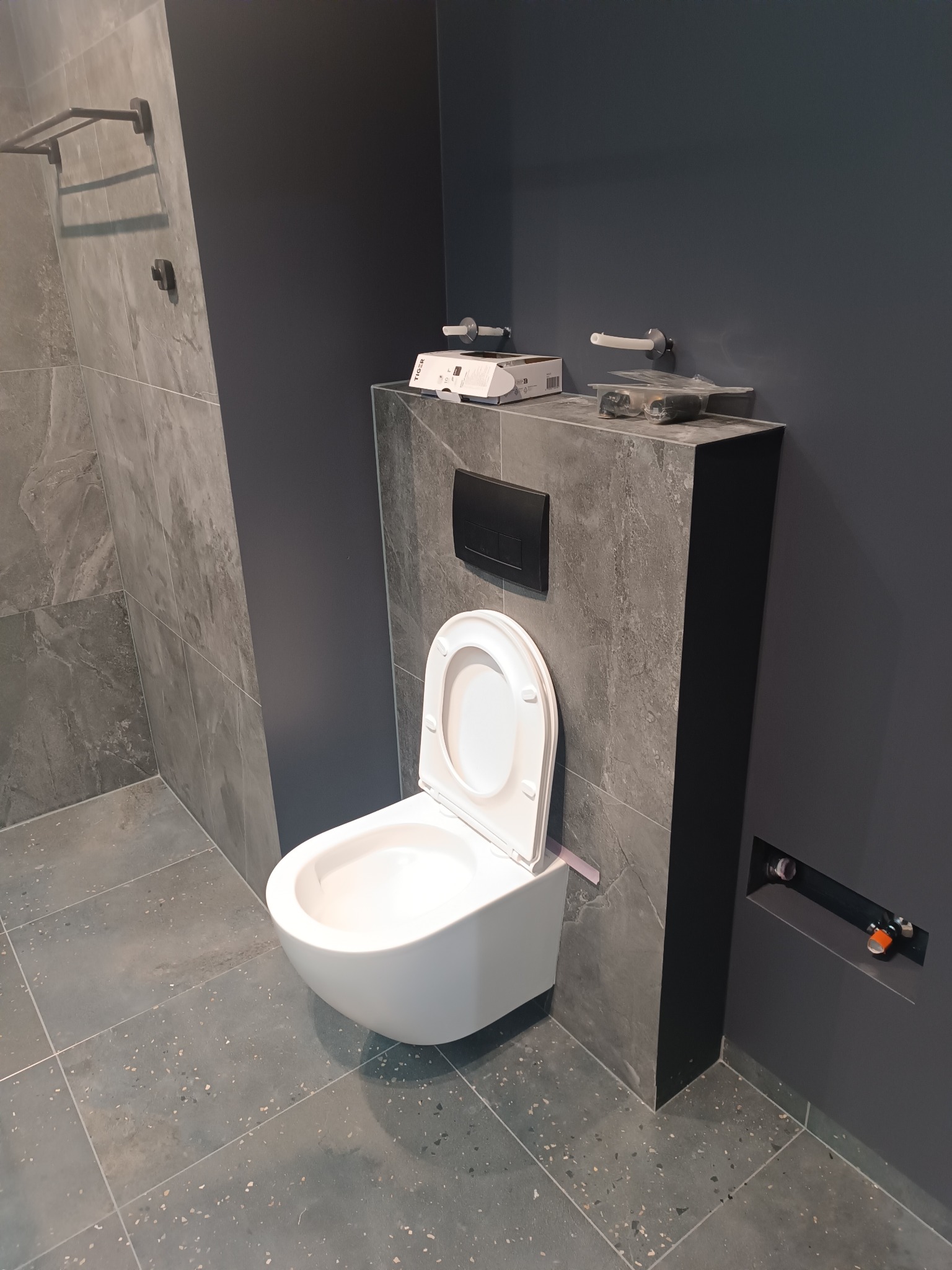 Nowoczesna, szara łazienka z podwieszaną toaletą. Widoczne płytki imitujące kamień na ścianach i podłodze, oraz czarne akcenty w postaci przycisku spłukującego.