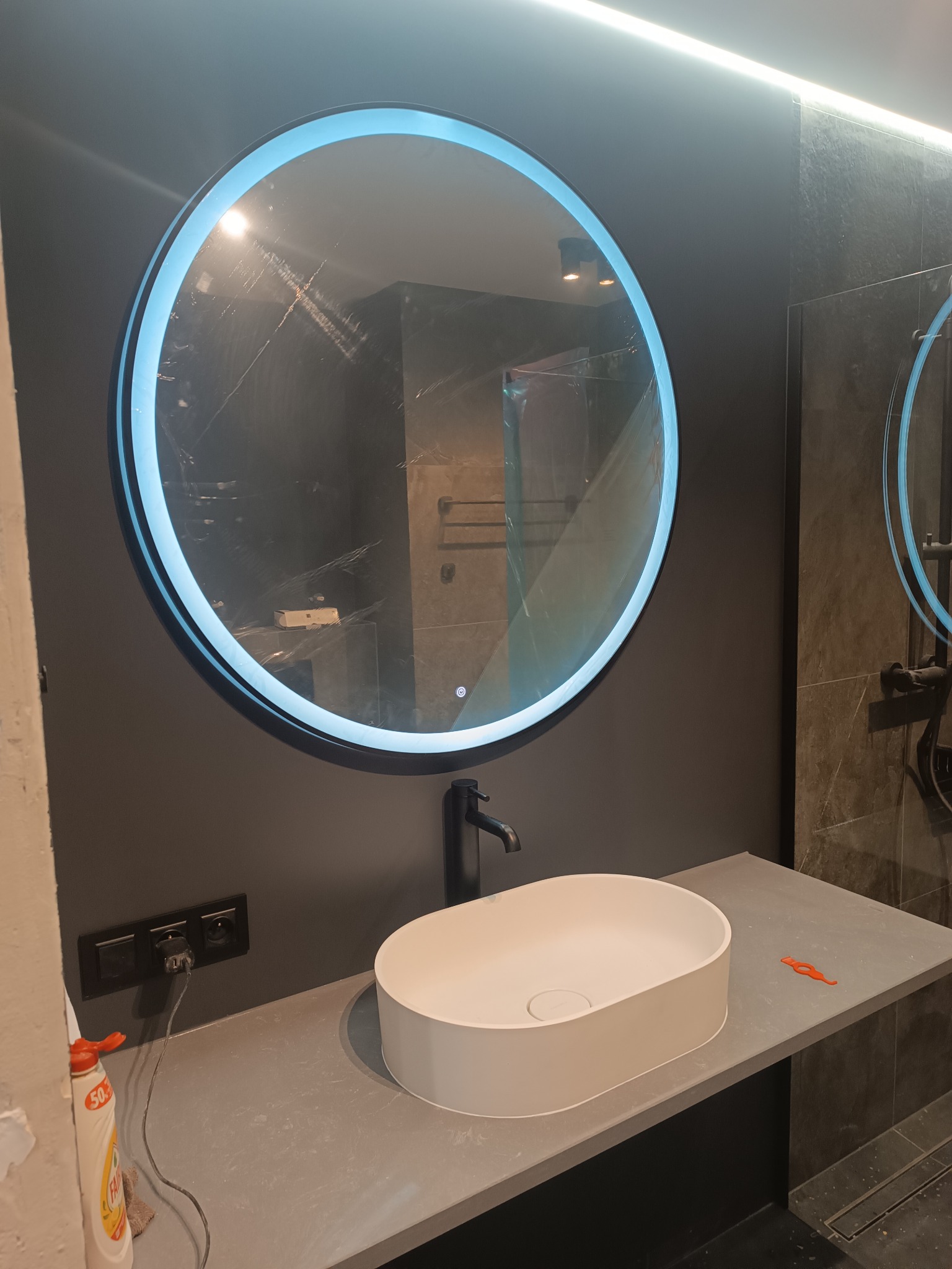 Nowoczesna łazienka z okrągłym lustrem LED, białą umywalką na szarym blacie i ciemnymi płytkami. Widoczne gniazdka i akcesoria, sugerujące etap wykończenia.