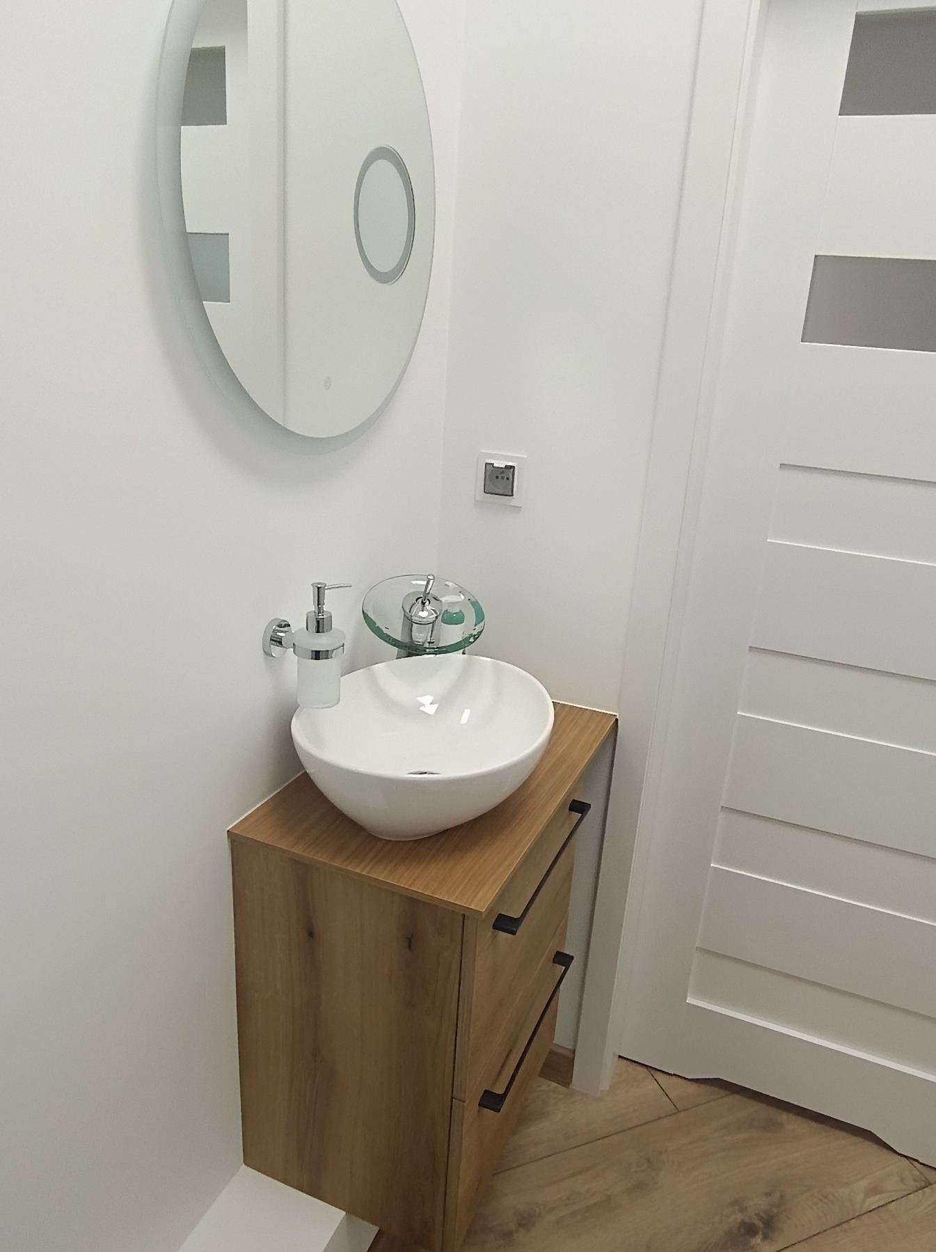 Nowoczesna łazienka: biała umywalka nablatowa na drewnianej szafce z czarnymi uchwytami, okrągłe lustro z podświetleniem i szklana bateria.