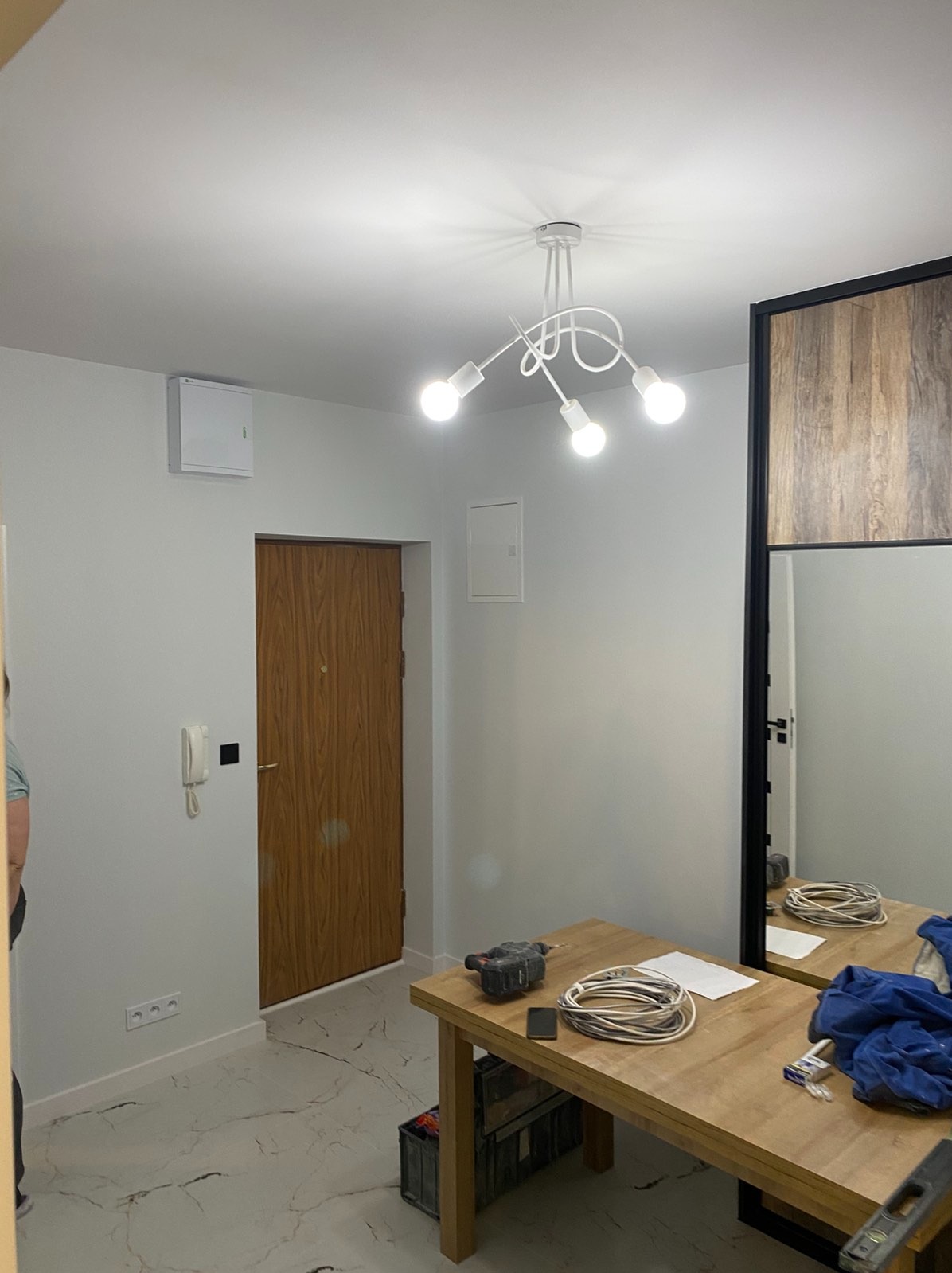 Jasne wnętrze po remoncie: białe ściany, marmurowe płytki podłogowe, drewniane drzwi i nowoczesna lampa sufitowa. Na stole narzędzia i kable.