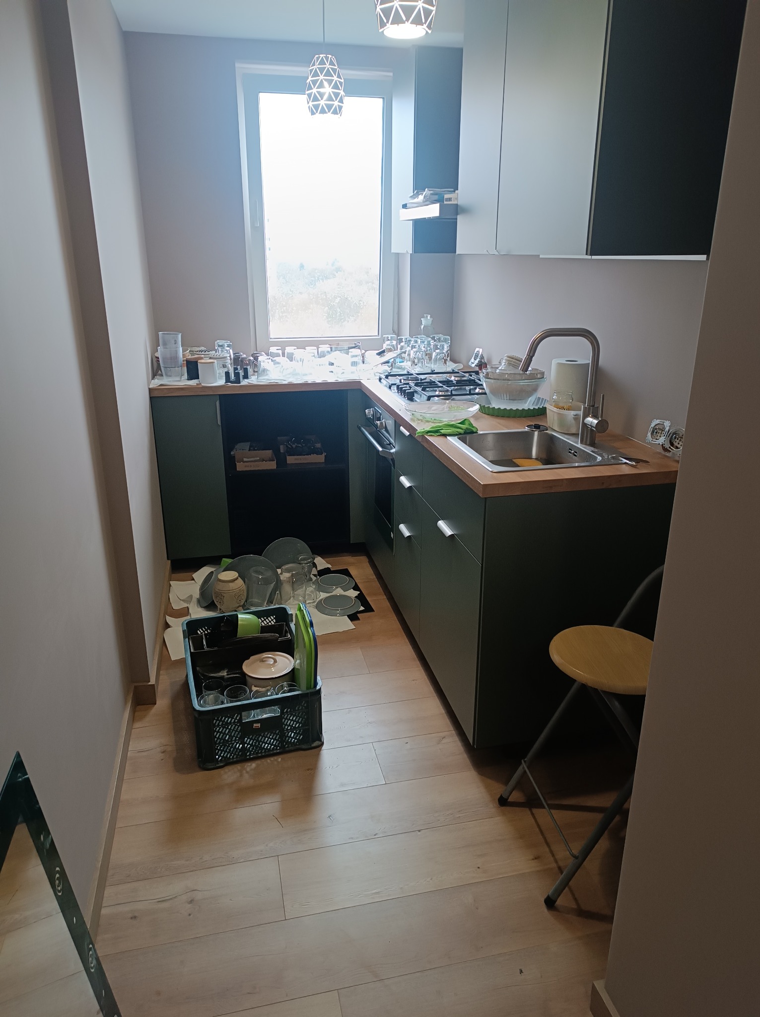 Umyte naczynia schnące w kuchni z zielonymi szafkami i drewnianym blatem. Widoczne szklanki, talerze i sztućce. Na podłodze skrzynka z naczyniami.