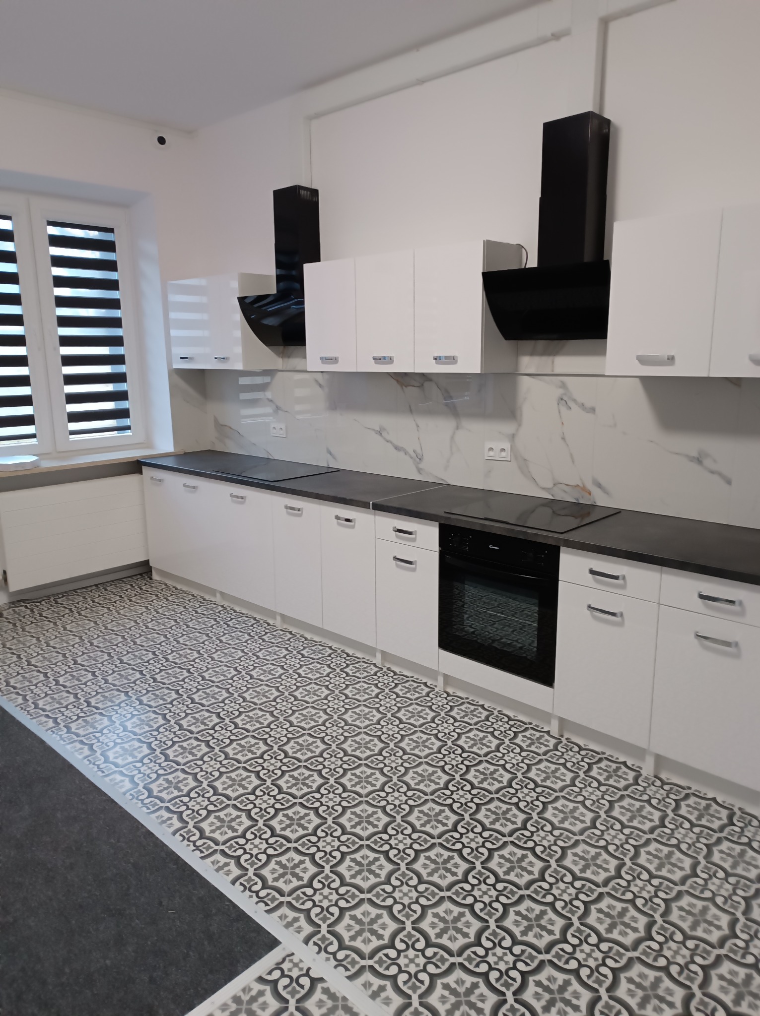 Nowoczesna kuchnia z białymi szafkami, czarnym blatem i wzorzystą podłogą. Okap w kolorze czarnym. Żaluzje w oknie. Minimalistyczny design.