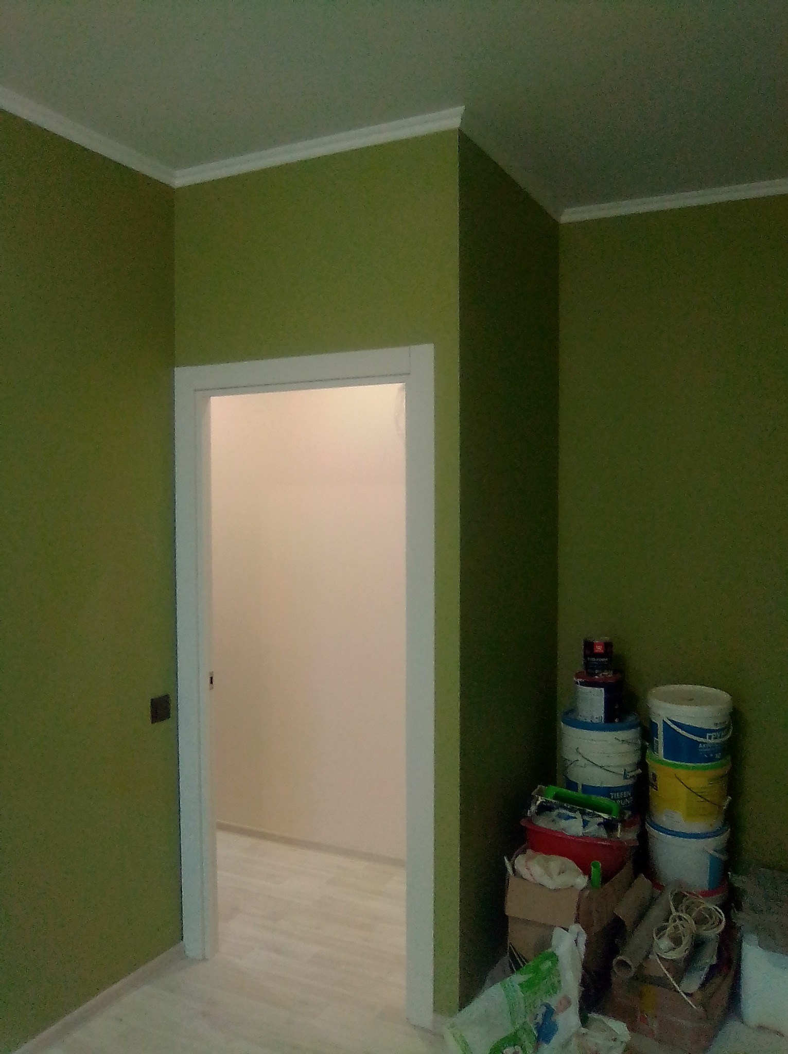 Świeżo pomalowane na oliwkowo ściany z białym wykończeniem sufitu i ościeżnicy. W tle widoczne białe ściany i podłoga. W rogu sterta wiader z farbą i narzędzi malarskich.