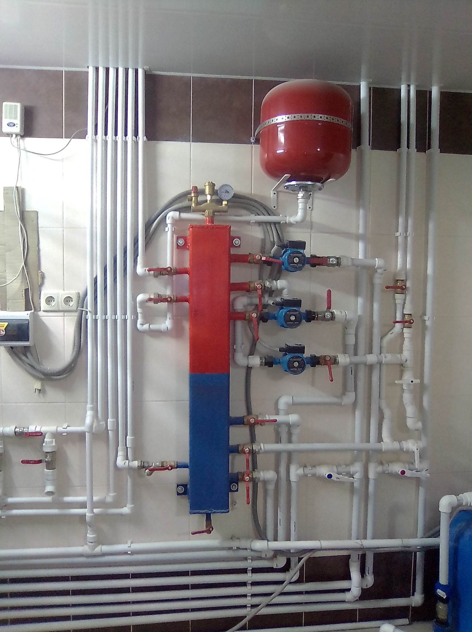 Instalacja centralnego ogrzewania z czerwonym zbiornikiem wyrównawczym i rozdzielaczem pomalowanym na czerwono i niebiesko, zamontowana na ścianie z płytkami.