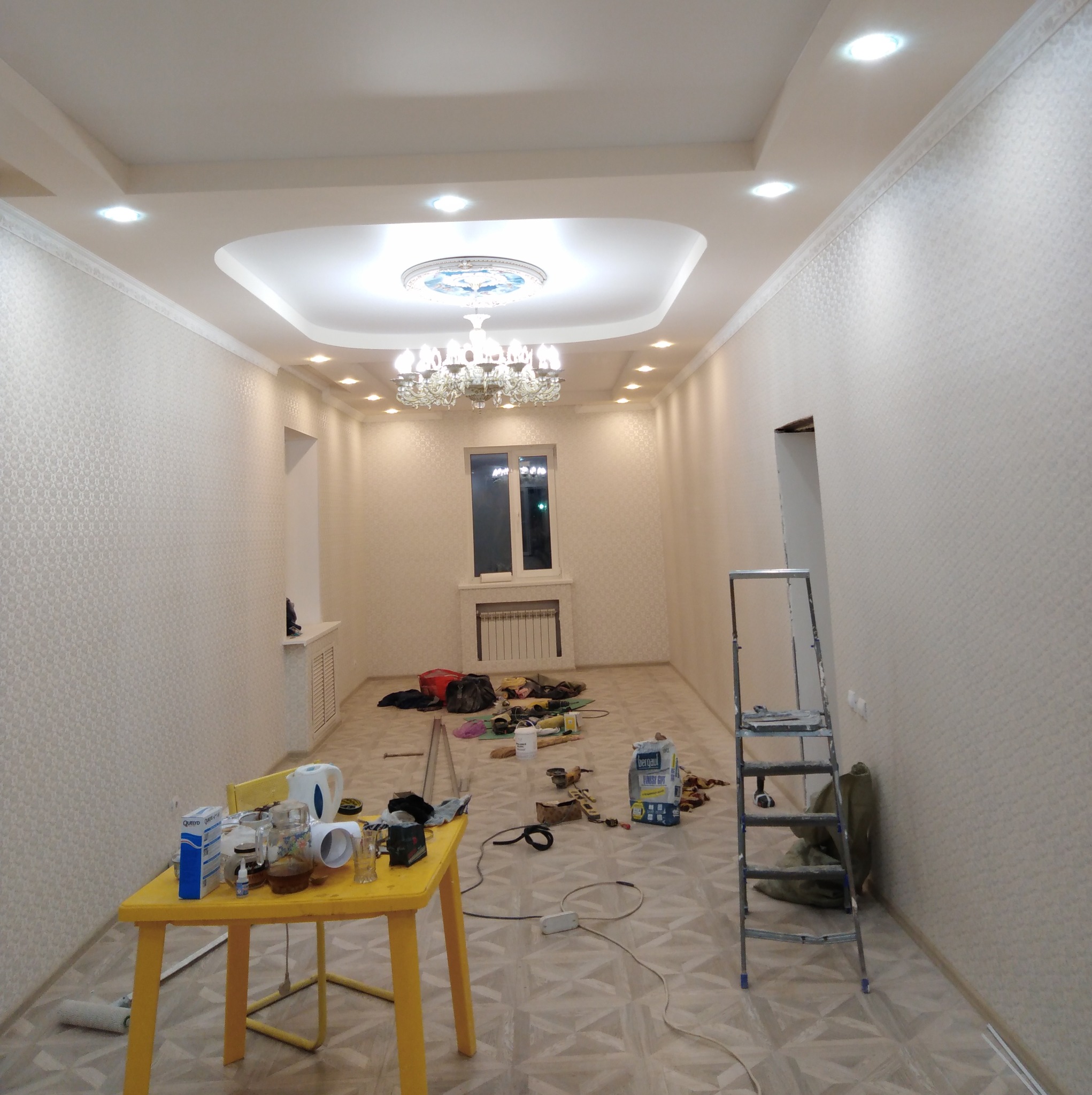 Pokój po remoncie: jasne ściany z tapetą, ozdobny sufit z żyrandolem, narzędzia na stole i drabina. Widok na okno i grzejnik. Etap wykończenia wnętrza.